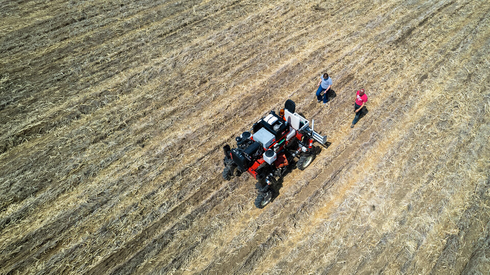 Aerial view of two people standing near a red tractor in a harvested field.