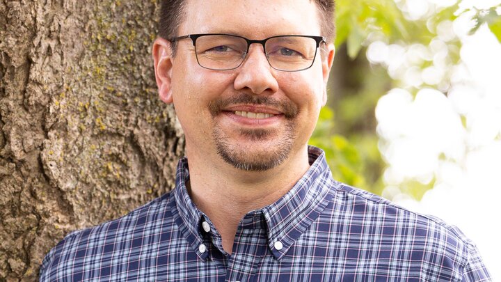 man smiling in front of a tree wearing a blue plaid shirt
