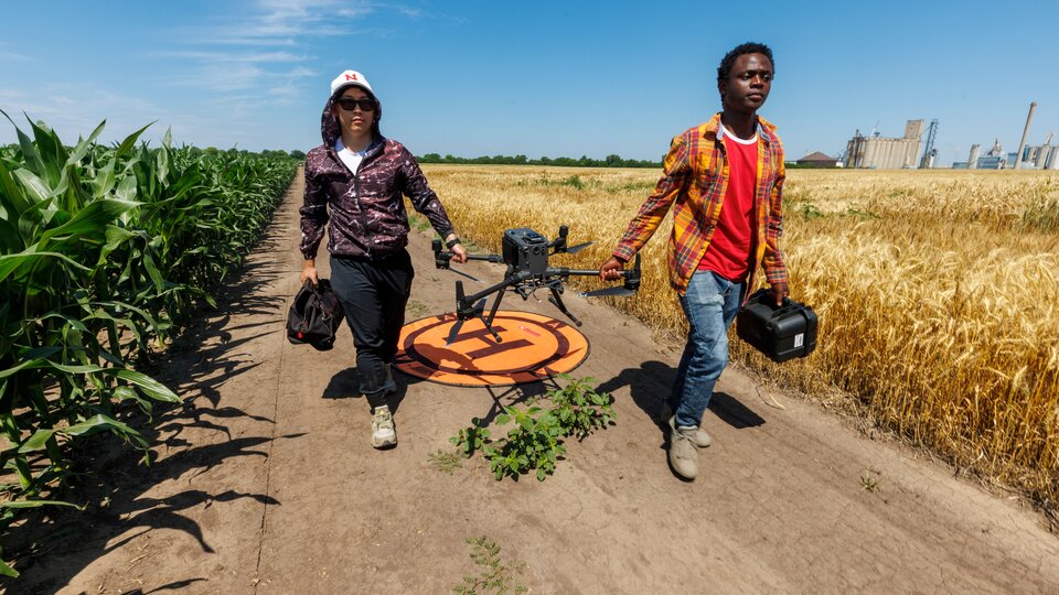 two students carrying  a drone in the middle of a field