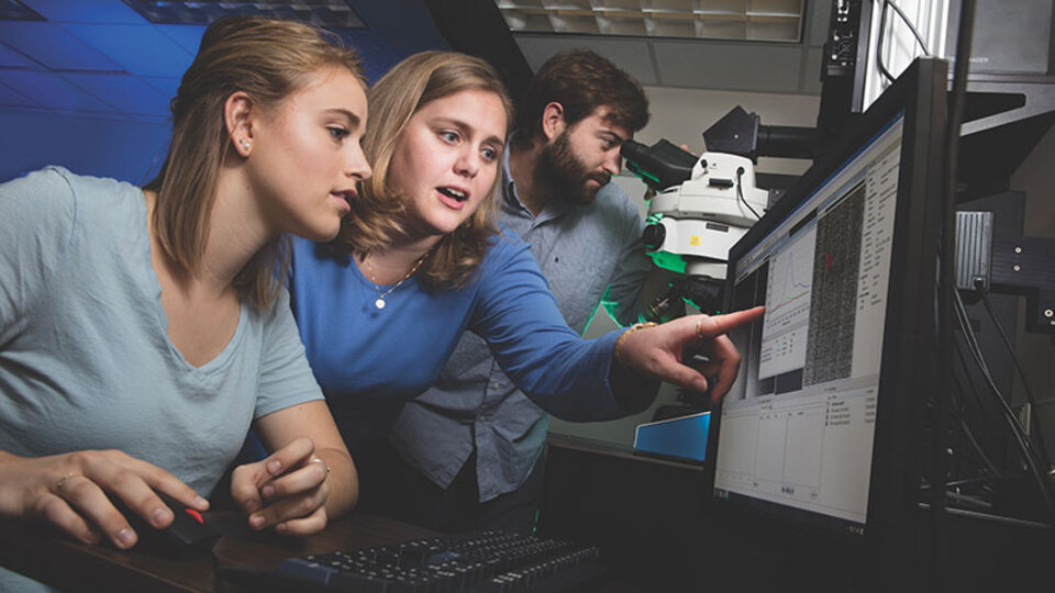 Image of three people in the lab. Dr.Iverson in the middle is pointing at a computer