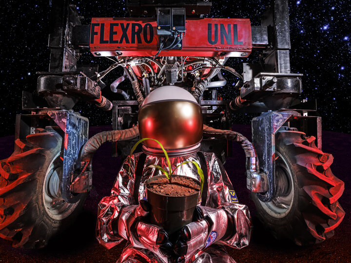 Man in a spacesuit holding vase with corn with a Flexrow UNL planter in the background